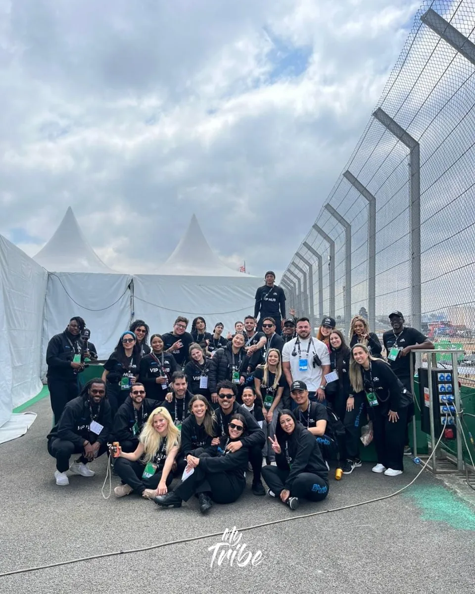 Equipe grande de staff da My Tribe com uniforme preto do VIP Hub, reunida ao lado das tendas do evento
