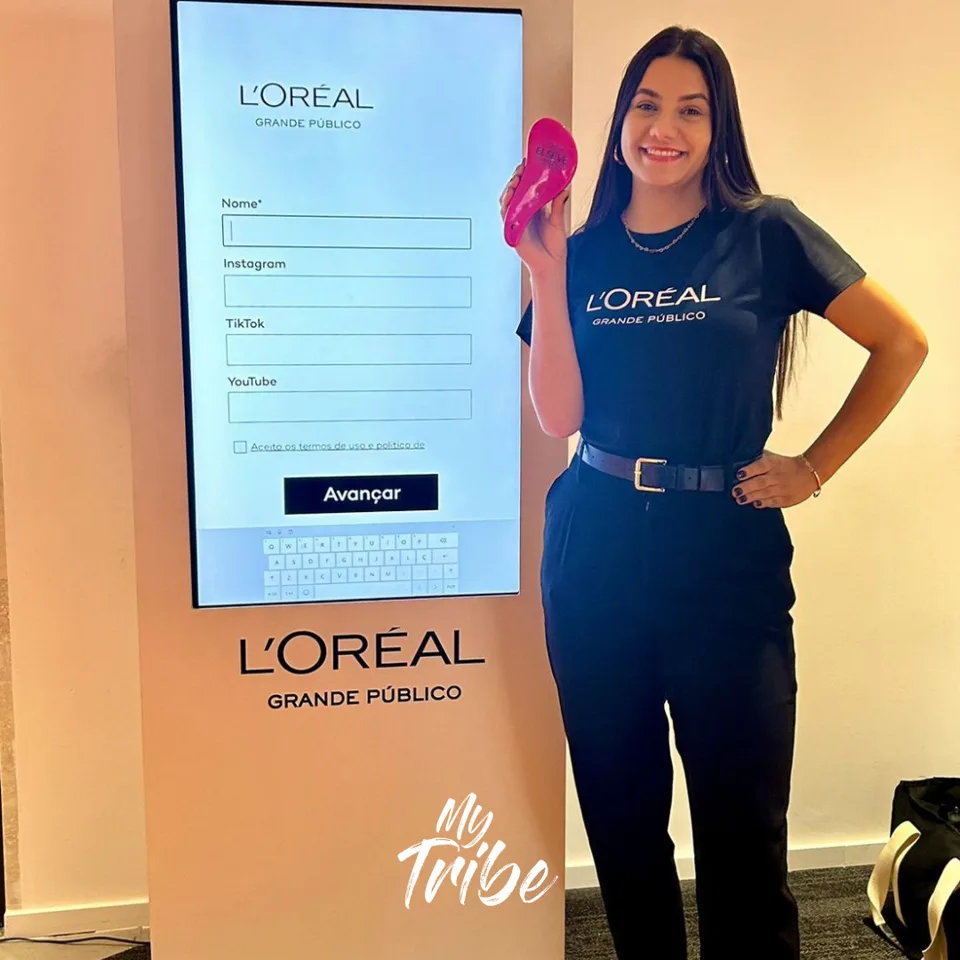Promotora segurando brinde ao lado de totem touch de cadastro da L'Oréal